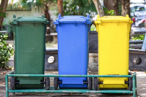 Three wheeled rubbish bins are lined up in a row on a paved outdoor area, with the left bin being green, the middle bin blue, and the right bin yellow. Each bin is made of durable plastic with a smooth matte finish and features a hinged lid on top, with the green and blue bins having slightly darker lids than their body. The bins are positioned in front of a low metal barrier, which has a horizontal bar supported by vertical posts and runs parallel to the bins, suggesting an organized waste collection point. Behind the bins, there is a background of blurred greenery, including trees and plants, indicating an outdoor setting, possibly on a residential or commercial premises in Acton. The natural lighting highlights the vibrant colors of the bins and the textures of the plastic surfaces, aligning with the services offered by Commercial Waste Removal Acton for rubbish disposal and waste management needs in the local area.