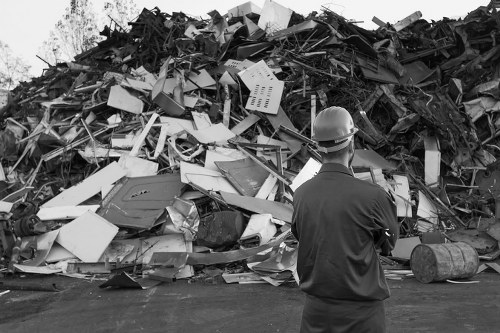 A man wearing a safety helmet and dark work clothing stands with his back to the camera, observing a large heap of mixed discarded waste materials on an outdoor surface. The waste includes various broken and discarded appliances, panels, and structural debris made of metal, plastic, and wood, some with visible textures such as smooth plastic and rough, splintered wood. The pile appears chaotic, with items leaning, stacked, and scattered in different directions, and the background shows an open environment with natural light. The scene relates to waste disposal or rubbish removal activities, consistent with services offered by Commercial Waste Removal Acton, serving the local area around W3 postal code.