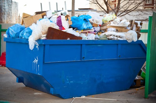 A large, blue industrial skip overflowing with mixed waste materials, including cardboard boxes, white and brown packaging, blue plastic bags, and crumpled paper, situated on a paved surface in an outdoor area. The waste extends over the top edges of the skip, with some items leaning against its sides. In the background, there are metal railings and the faint outline of buildings, suggesting a commercial or industrial environment, possibly within the Acton area. The skip's surface shows some signs of use, with minor scratches and scuffs, and the overall scene indicates waste collection or rubbish removal in progress. The image is associated with services provided by Commercial Waste Removal Acton, focusing on effective rubbish clearance for local businesses or premises.