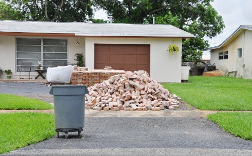 A residential driveway in front of a single-storey house with a white exterior and a brown garage door, featuring a large pile of light-colored bricks and rubble positioned on the asphalt surface. To the left of the pile, there is a dark grey wheeled rubbish bin with a white plastic bag or cover placed on top. The house has a large window with horizontal blinds and a small decorative plant on the left side, while the surrounding area includes a well-maintained green lawn and mature trees providing shade. In the background, the house's roof is visible, along with another building on the right side, indicating a suburban setting. This scene visually represents a typical rubbish removal or waste clearance process, performed by services such as Commercial Waste Removal Acton, to clear construction debris or renovation waste from residential properties in the local area.