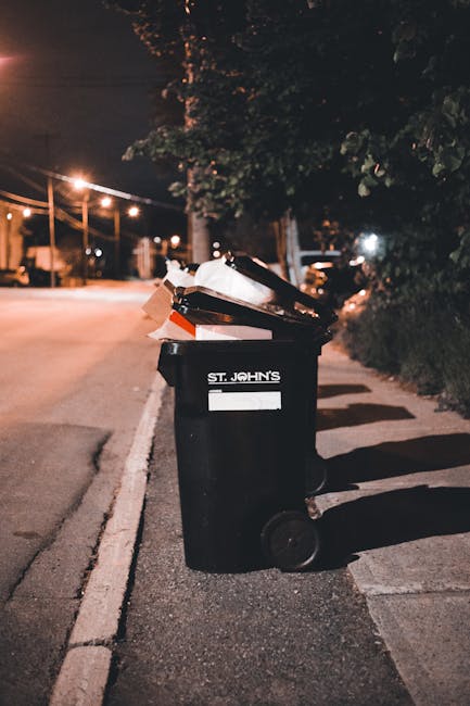 A row of large, rectangular wheelie bins positioned along a pavement beside a red brick wall. The bins are made of durable plastic, with textured lids featuring raised ridges for grip, and are painted