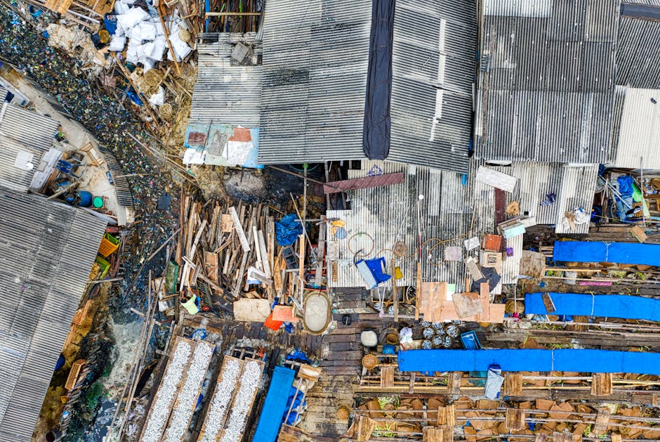 A large outdoor waste collection point featuring multiple overflowing rubbish bins and discarded debris on the ground in front of a commercial area. The central metal bin labeled for mixed paper and c